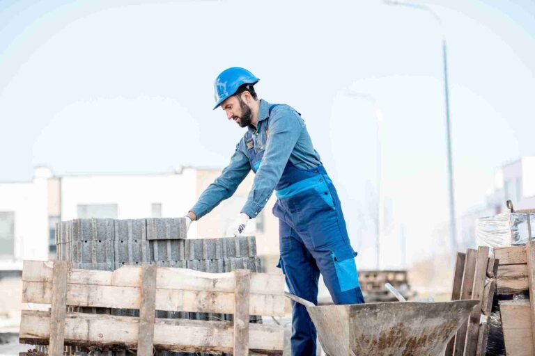 Construction worker wearing a blue hard hat and gloves stacking concrete paving blocks on a pallet at an outdoor job site, with a wheelbarrow nearby.